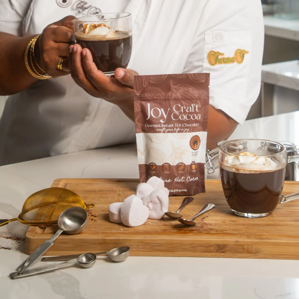 A chef in a white uniform holding a glass mug of Joy Craft Cocoa with toasted marshmallows, next to a second mug, a product pouch, and heart-shaped marshmallows on a wooden board.