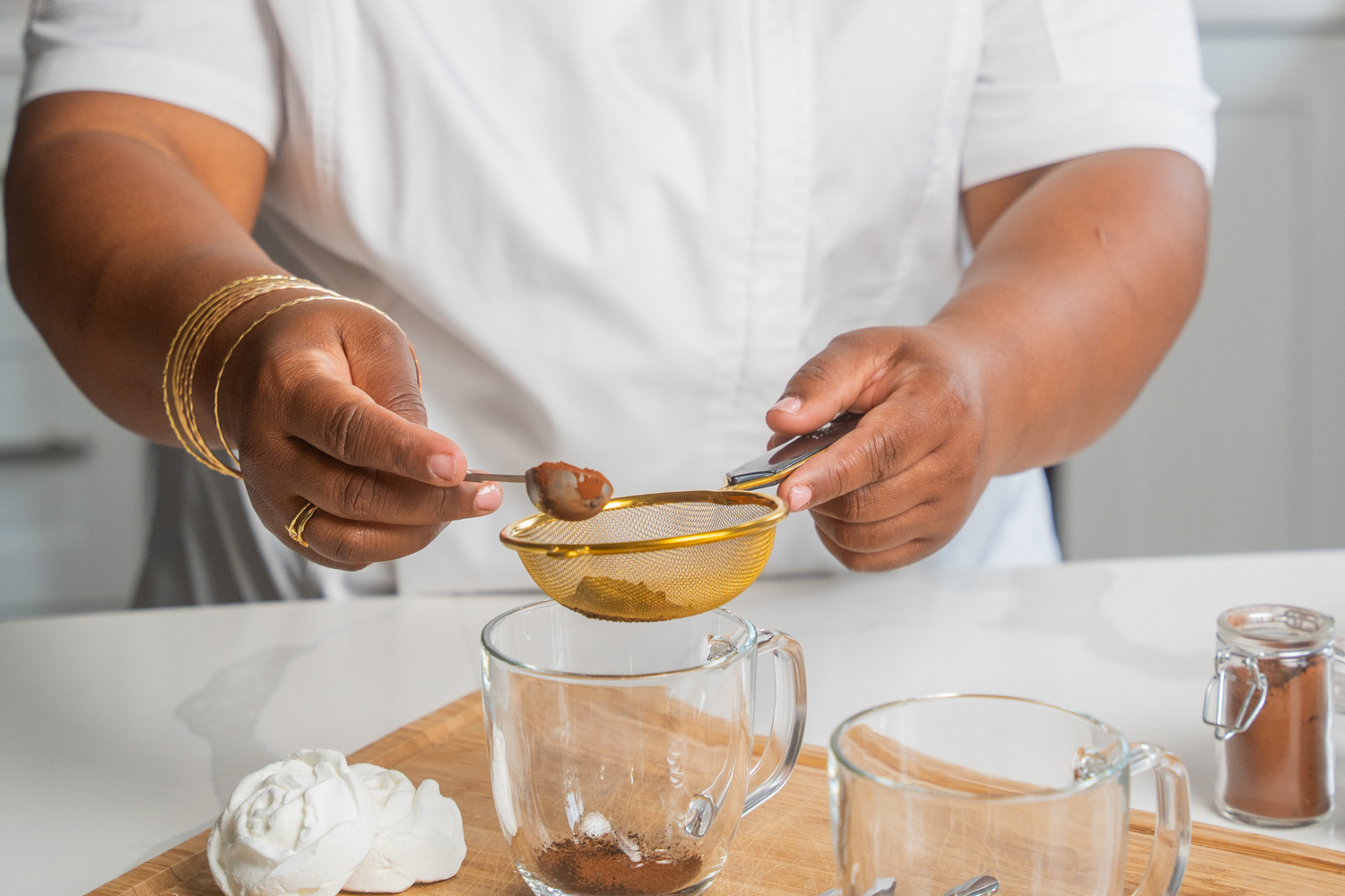 Person sifting a powdery substance into a glass mug on a wooden board.